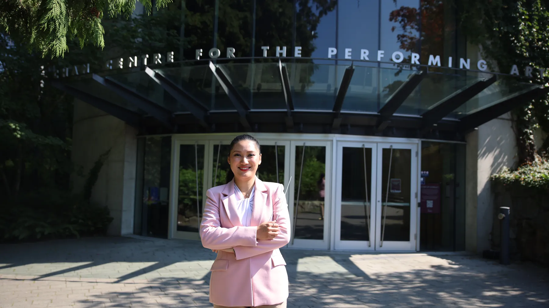 Meijun Chen stands in front of the Chan Centre for the Performing Arts wearing a pink blazer and holding a conductor's baton.