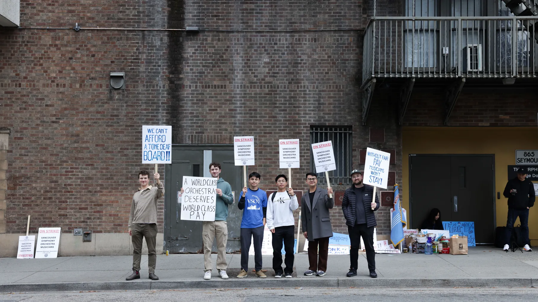 A line of VSO musicians stand outside the stage door of the Orpheum, holding signs that read "We can't afford a tone deaf board!" and "On strike!"
