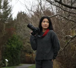 Cassidy Chen, a young woman with dark hair, wears a grey sweater and red scarf, and stands in front of autumn trees. She holds up a camera and looks to the side, subtly smiling.