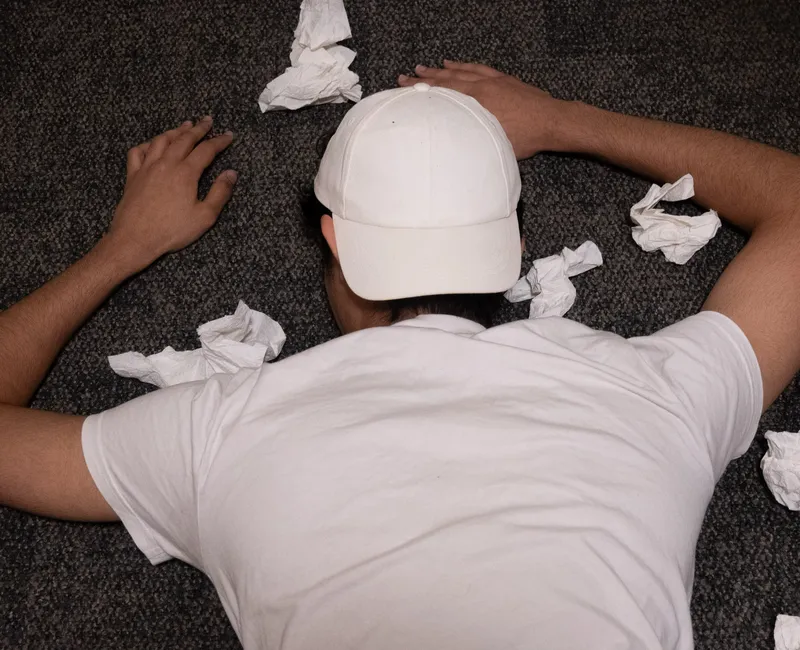 A person lies on the floor wearing a backwards baseball cap, surrounded by balled-up tissues.