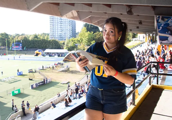 A photo of a girl holding a book in front of a football field wearing a UBC Thunderbirds jersey.