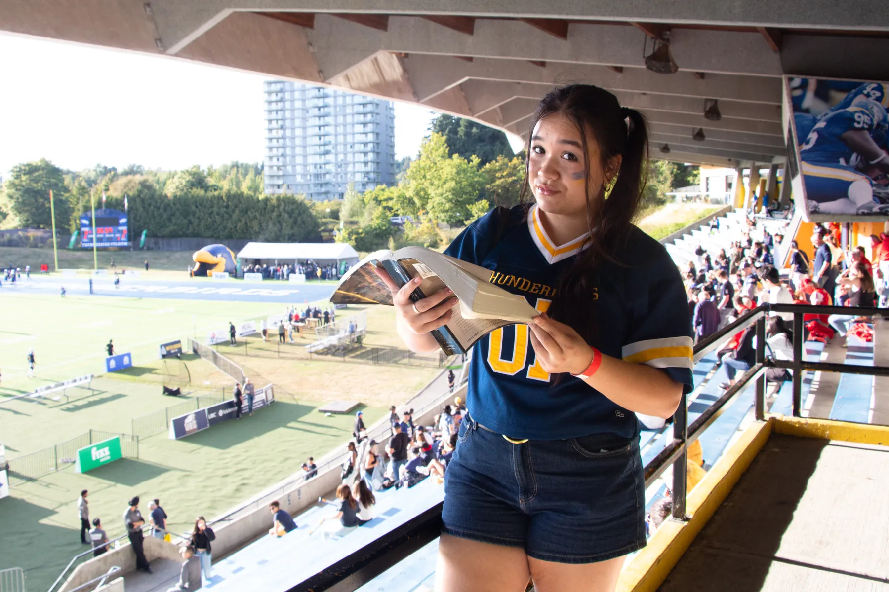 A photo of a girl holding a book in front of a football field wearing a UBC Thunderbirds jersey.
