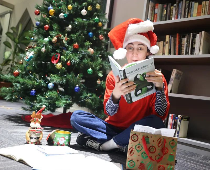 A smiling person wearing a Santa hat sits in front of a Christmas tree and reads an academic book.