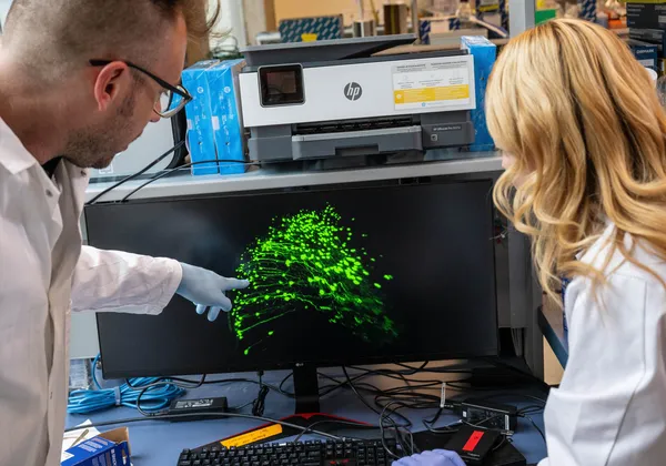 Two scientists in lab coats examine a computer screen displaying a glowing green network of neurons. One of them points to the display.