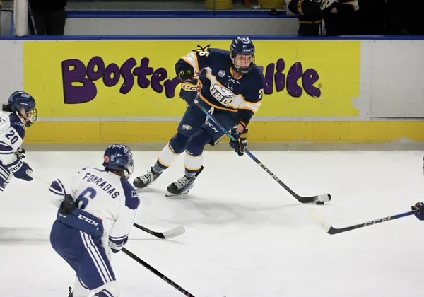 A hockey player in a navy blue jersey kit and helmet is centred in frame, with the puck on her stick in front of a board ad for Booster Juice. Two players in white jerseys and light blue pants are racing toward the player in the navy blue player.