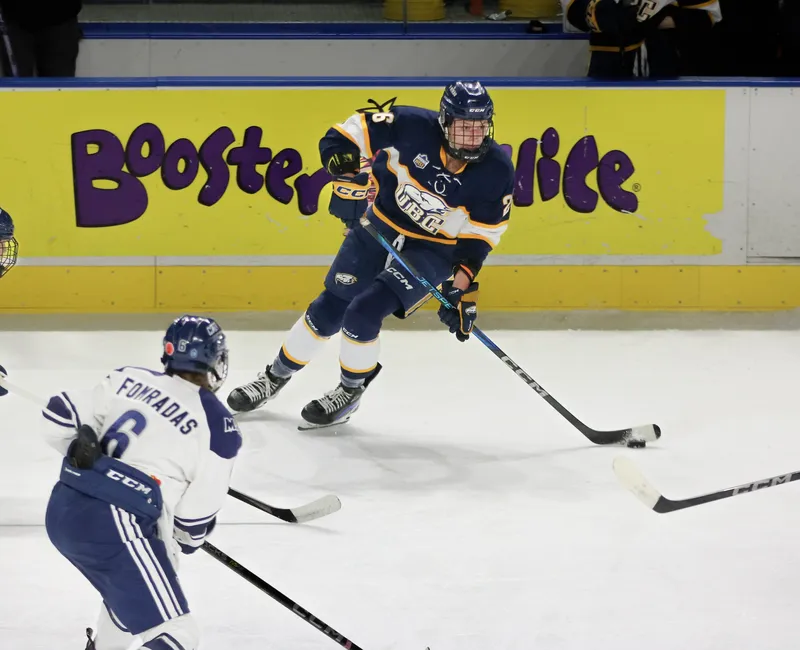 A hockey player in a navy blue jersey kit and helmet is centred in frame, with the puck on her stick in front of a board ad for Booster Juice. Two players in white jerseys and light blue pants are racing toward the player in the navy blue player.