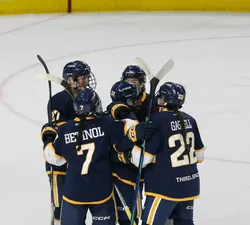 Five female hockey players in navy blue jerseys huddle on the ice in celebration of a goal.