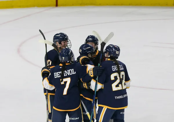 Five female hockey players in navy blue jerseys huddle on the ice in celebration of a goal.