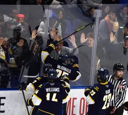A hockey player in a blue jersey jumps into the plexiglass in celebration while spectators cheer and hit the plexiglass.