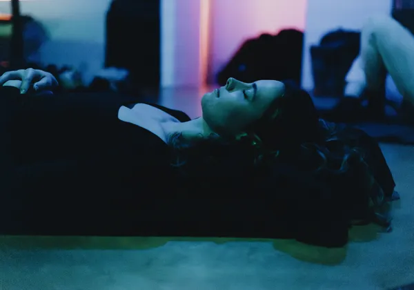 In dim green and pink lighting, a young woman lies on the ground with her eyes shut.