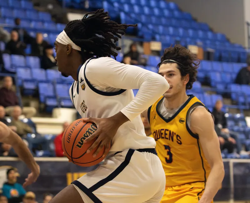 A male basketball player in a white jersey kit holds the ball in his hands, driving to the net.