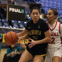 A woman basketball player in a green "Alberta" jersey drives to the hoop with the ball in her hand.