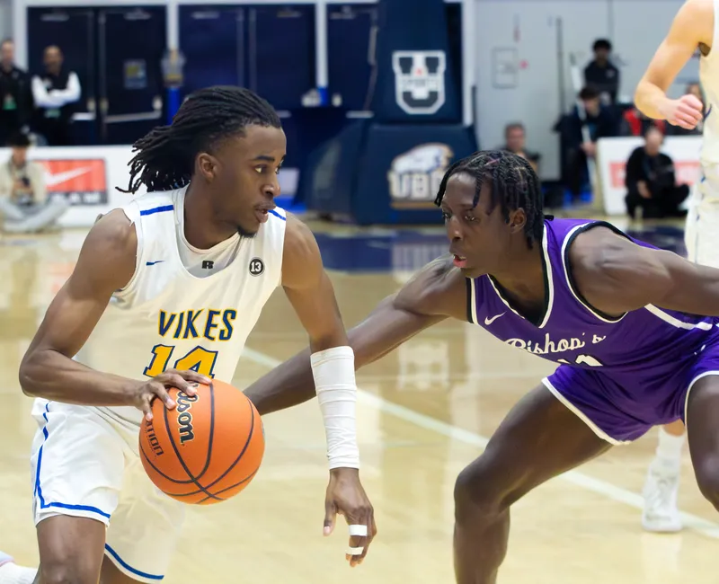 A Black male basketball player in a white "Vikes" jersey dribbles the ball while being defended by a Blak basketball player in a purple "Bishop's" jersey.