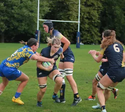 A female rugby player with blonde hair is carrying the ball in a navy blue jersey kit. She is trying to break through a tackle made by another rugby player, wearing a light blue and yellow jersey kit. Three other players in navy blue jerseys are in the frame, facing away from the camera. The background is a lush green outdoor rugby pitch, with the goalposts in the centre of the photo.