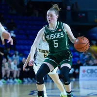 A woman basketball player in a green jersey kit that says "Huskies 5" dribbles with the ball.