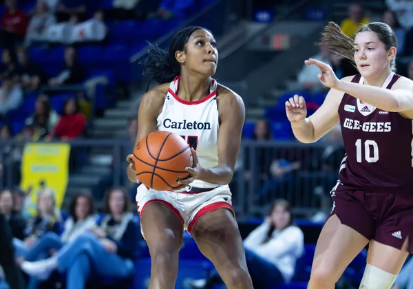 A woman basketball player in a white "Carleton" jersey holds the ball mid-play.