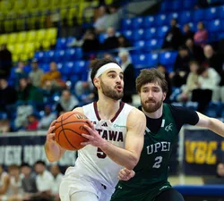 A man basketball player in a white jersey kit and a white headband drives with the ball in both hands while another player in a green jersey chases after him.
