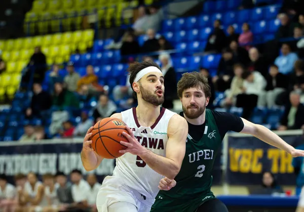 A man basketball player in a white jersey kit and a white headband drives with the ball in both hands while another player in a green jersey chases after him.