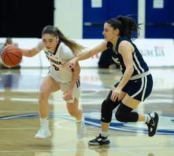 A woman basketball player in a white jersey kit dribbles with the ball while another woman player struggles to defend.