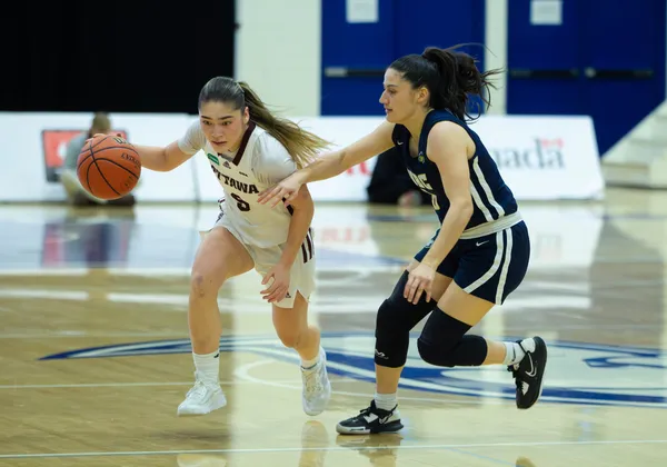 A woman basketball player in a white jersey kit dribbles with the ball while another woman player struggles to defend.