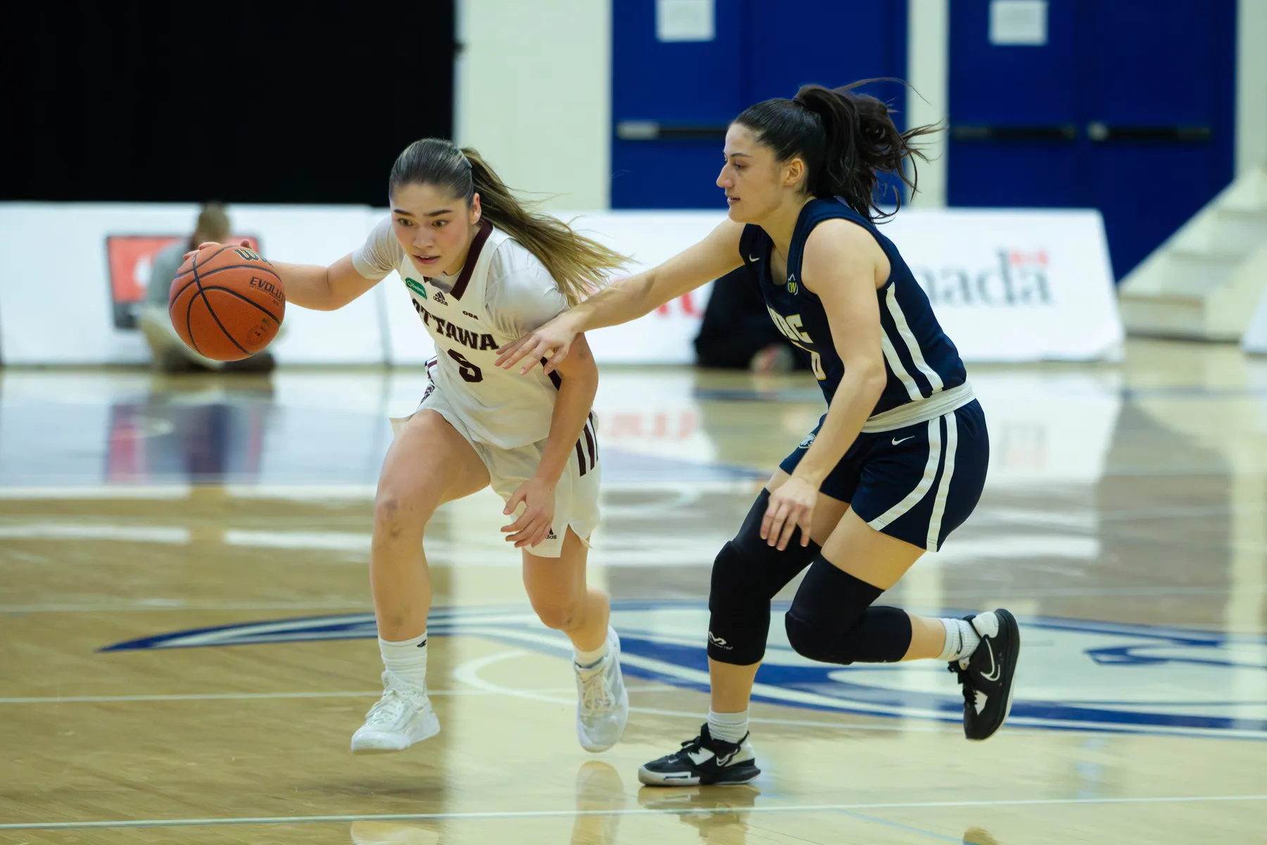 A woman basketball player in a white jersey kit dribbles with the ball while another woman player struggles to defend.