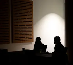 Two dark figures are silhouetted against a bright spot of light on a white wall behind them.