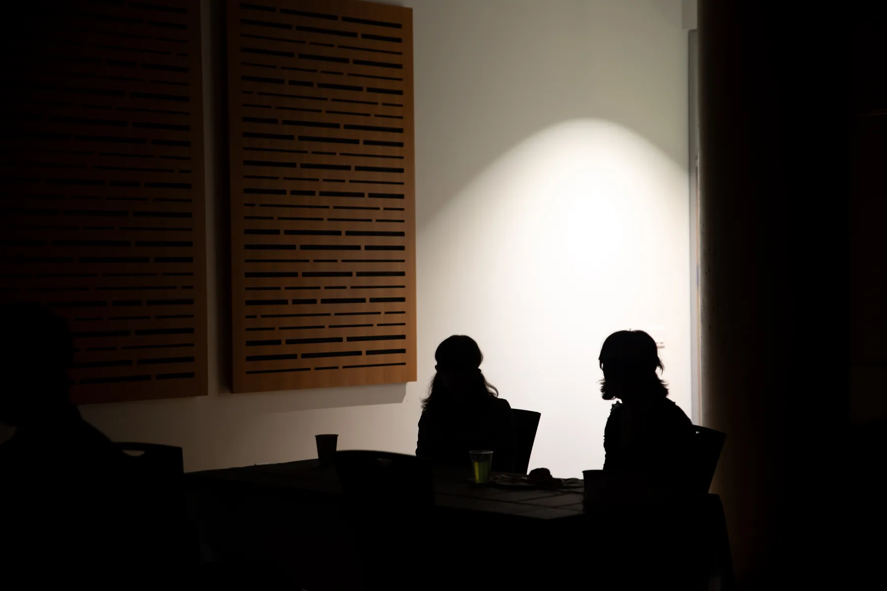 Two dark figures are silhouetted against a bright spot of light on a white wall behind them.