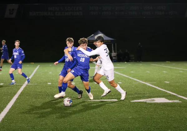 Two soccer players clash in pursuit of the ball on a football field at night. The player on the left is in a blue jersey kit that says "HORNS" on the front and the player on the right is in a white jersey kit.