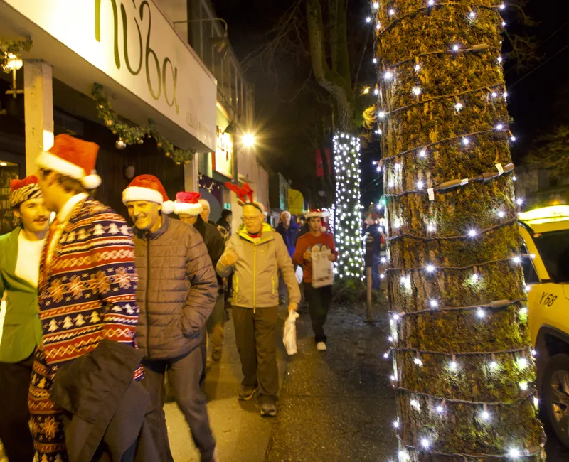 Men in Santa hats walk down a sidewalk with trees that have been decorated with lights. The men are slightly blurred from movement.