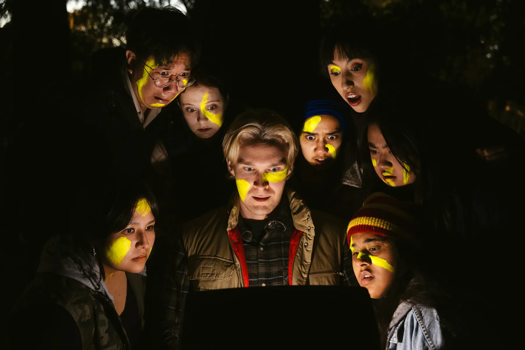 Eight cast members of Mr. Burns play, looking into a screen with yellow paint strikes on their faces.