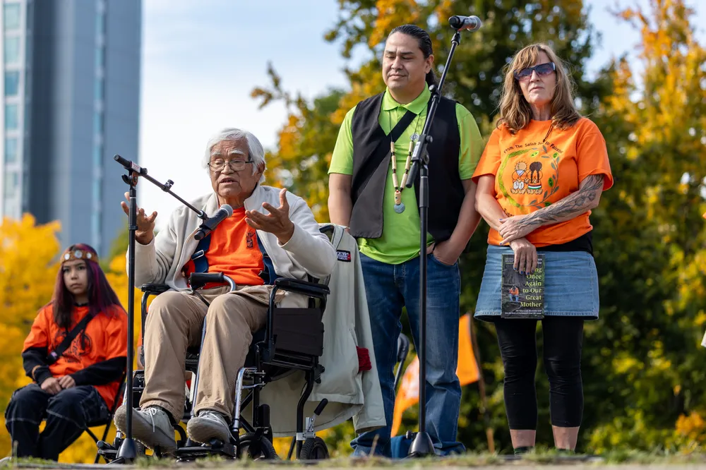 Elder Bill Jones addresses the crowd sitting in a wheelchair and gestures with his hands. Richard Wolff stands behind and to the right,