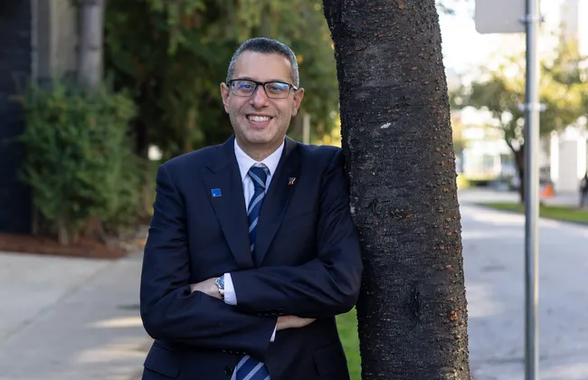 Kareem Allam leans against a tree. He is smiling and is wearing a suit and tie.