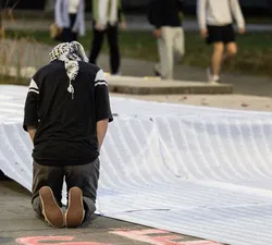 A person wearing a keffiyeh kneels in front of a long list of names.