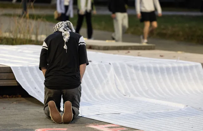 A person wearing a keffiyeh kneels in front of a long list of names.