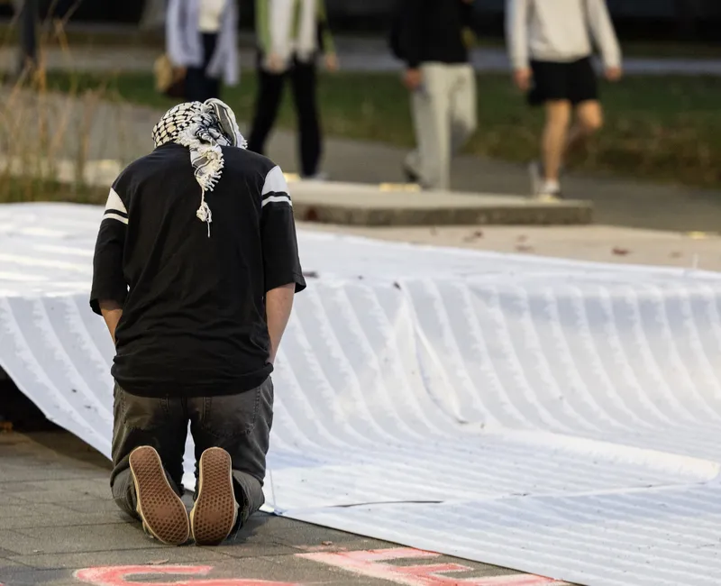 A person wearing a keffiyeh kneels in front of a long list of names.
