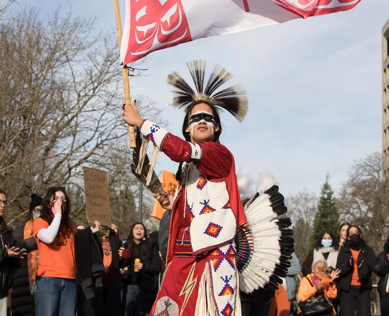 A person wearing traditional Indigenous regalia, including a feathered headdress, holds a red-and-white flag as they stand in front of a crowd at an outdoor demonstration. People behind them watch, clap, and hold signs among leafless trees and campus buildings.