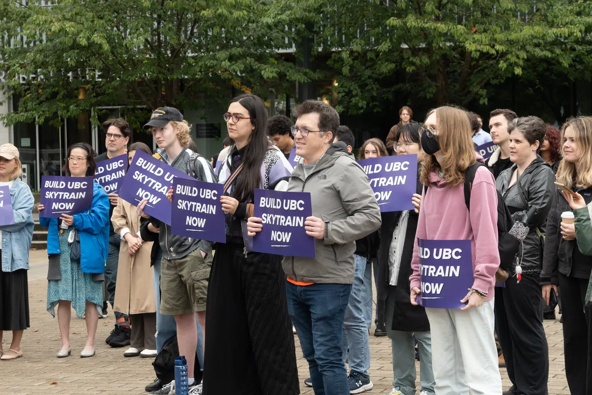 Students holding placards saying "Build UBC SkyTrain now" listen to speakers at the rally.