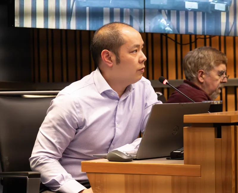 A man sits at the dais inside a meeting room.