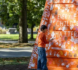 A photo of a child placing a hand print on the Engineering cairn, which was painted orange for the event and read: "Every Child Matters" and "Residential School denialism is a hate crime!"