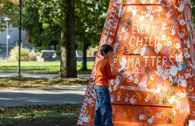 A photo of a child placing a hand print on the Engineering cairn, which was painted orange for the event and read: "Every Child Matters" and "Residential School denialism is a hate crime!"