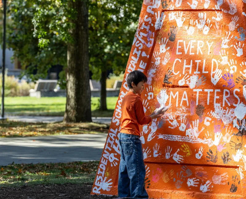 A photo of a child placing a hand print on the Engineering cairn, which was painted orange for the event and read: "Every Child Matters" and "Residential School denialism is a hate crime!"