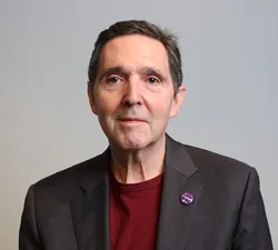 Photo of a man in a red shirt and dark grey blazer facing the camera with a grey background.