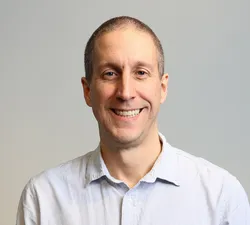 A man in an white collared shirt looking straight ahead with a white background.