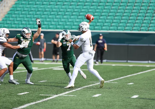 A football player in an all white uniform throws the football while players in green jerseys try to block it on a football field with empty stands.