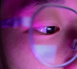 A close-up of someone's eye. In the reflection of their pupil is a tiny screen. The rest of their face is illuminated by magenta lighting.