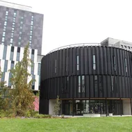 A large, round, black building sits in a grassy area. Pictured is the Arts Student Centre.