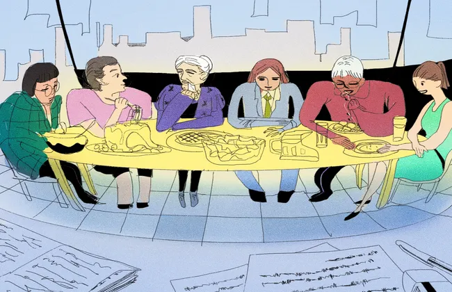 Group of people in office sitting around a long table eating a meal. In the foreground, pen and paper are laid on a table.