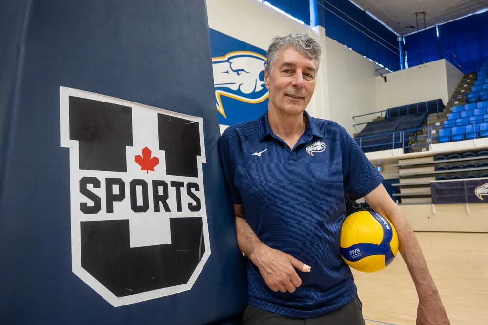 Reimer leans against a pillar with the U Sports logo on it, with a volleyball under his arm.