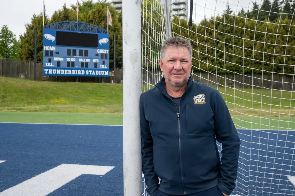 Mike Mosher leans against the goalpost at Thunderbird Stadium, with the scoreboard visible in the background.