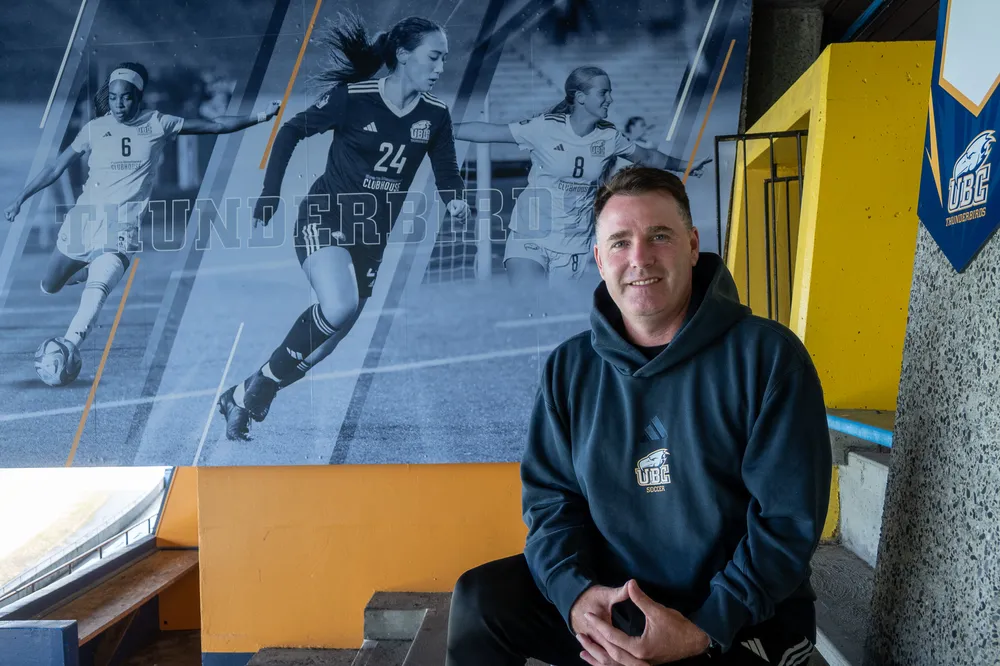 Jesse Symons sits in front of a mural of three of his current players at Thunderbird Stadium.